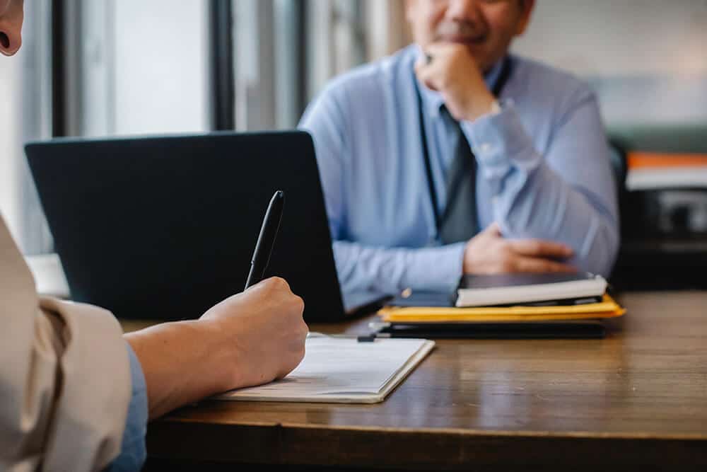 Person taking notes across from person on laptop